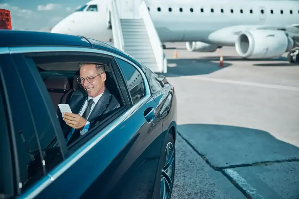 Professional chauffeur waiting in luxury car at Logan Airport terminal.
