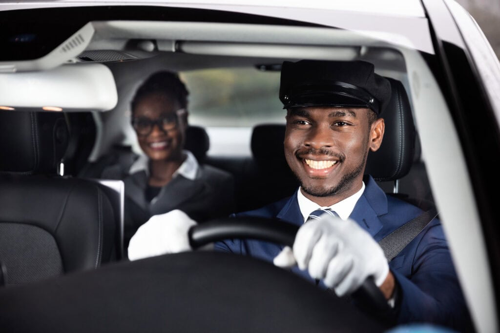 Smiling chauffeur driving a luxury car with a passenger in the back seat.