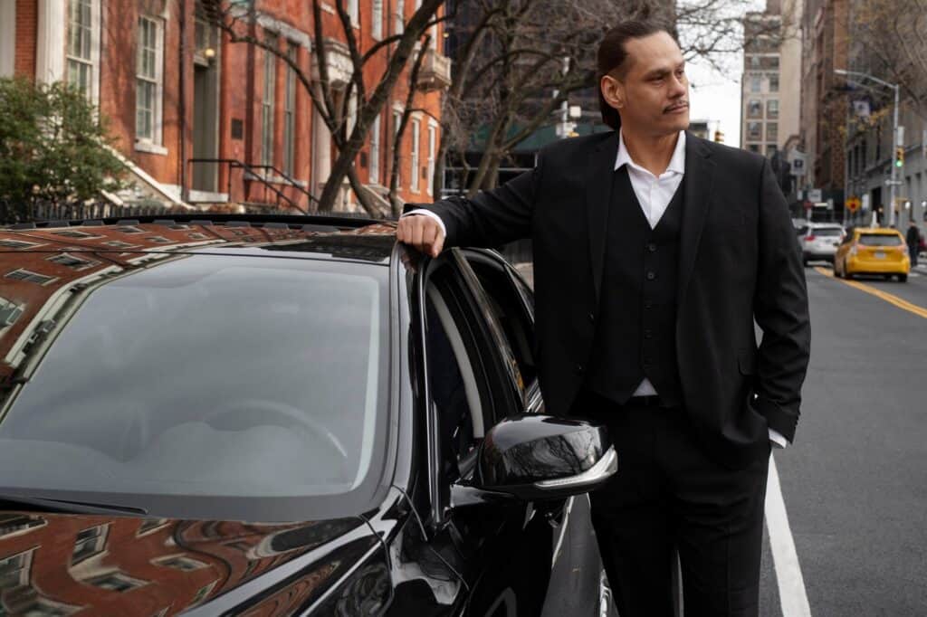 Professional chauffeur in a black suit leaning on a luxury car in NYC.