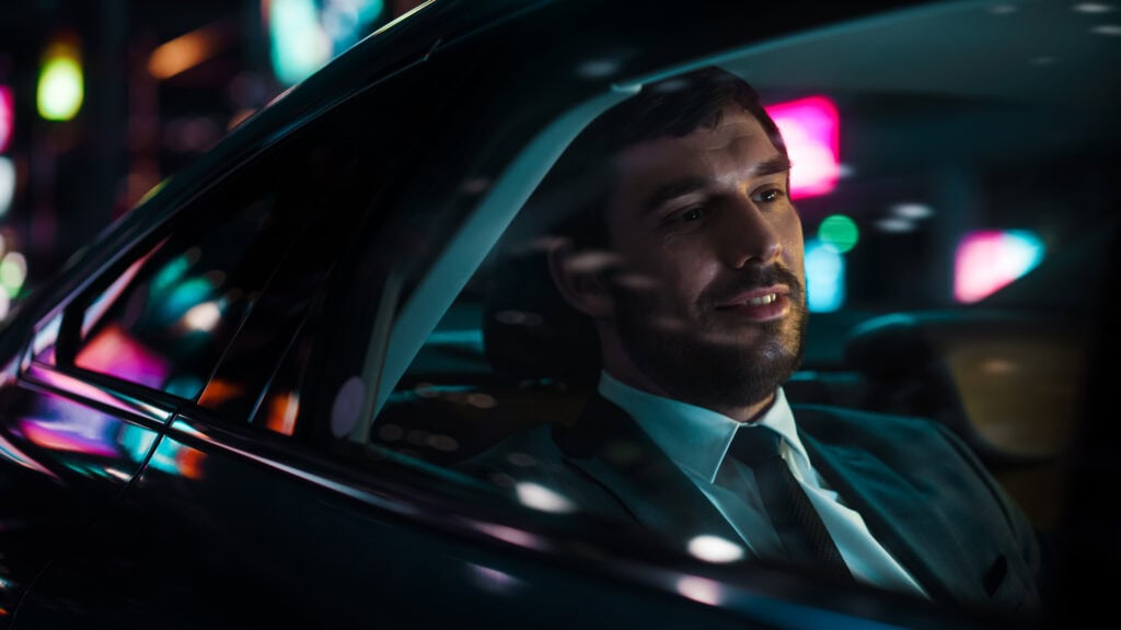 Man in suit driving luxury car at night with city lights in background.