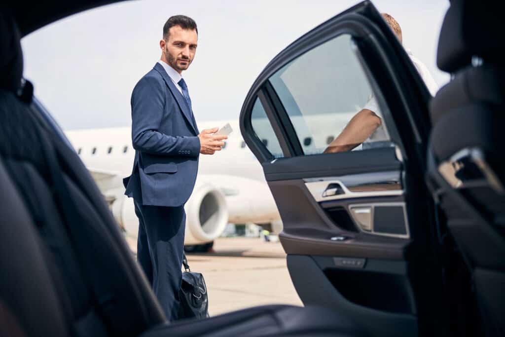 Chauffeur in suit waiting outside luxury car at airport.