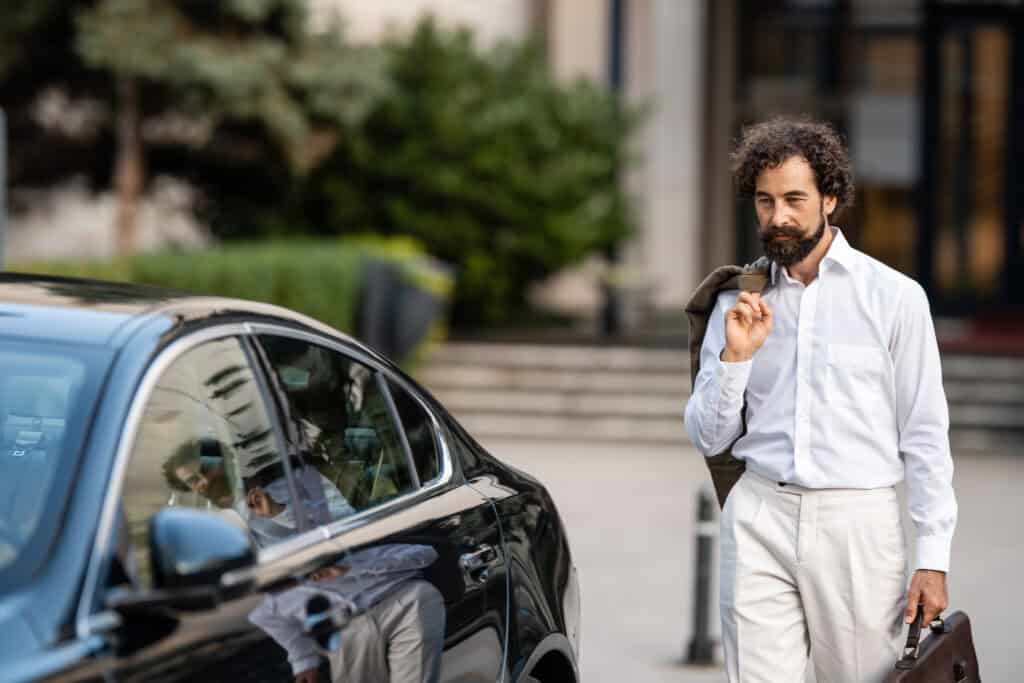Professional chauffeur with briefcase standing next to luxury car in urban setting.