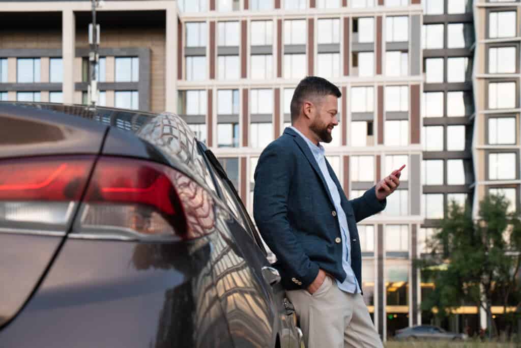 Man using smartphone next to luxury car in urban setting for chauffeur service.
